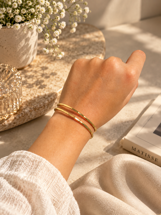 A close-up of a person's wrist wearing two slim gold bangle bracelets, styled with a linen sleeve, a vase of baby's breath, and a book in warm sunlight.