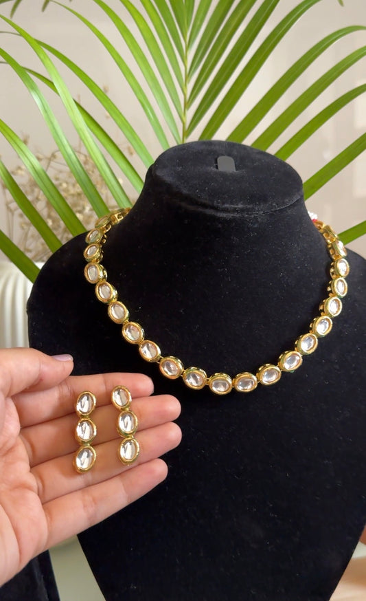 Gold-tone statement necklace and matching stud earrings displayed on a black jewelry bust, with a hand holding the earrings in front and green palm leaves in the background.