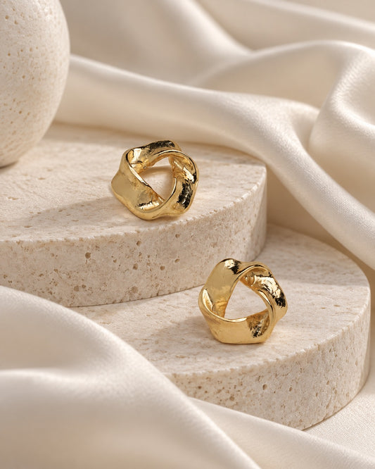 A pair of gold-toned twisted knot ring earrings displayed on round beige stone pedestals with draped cream satin fabric.