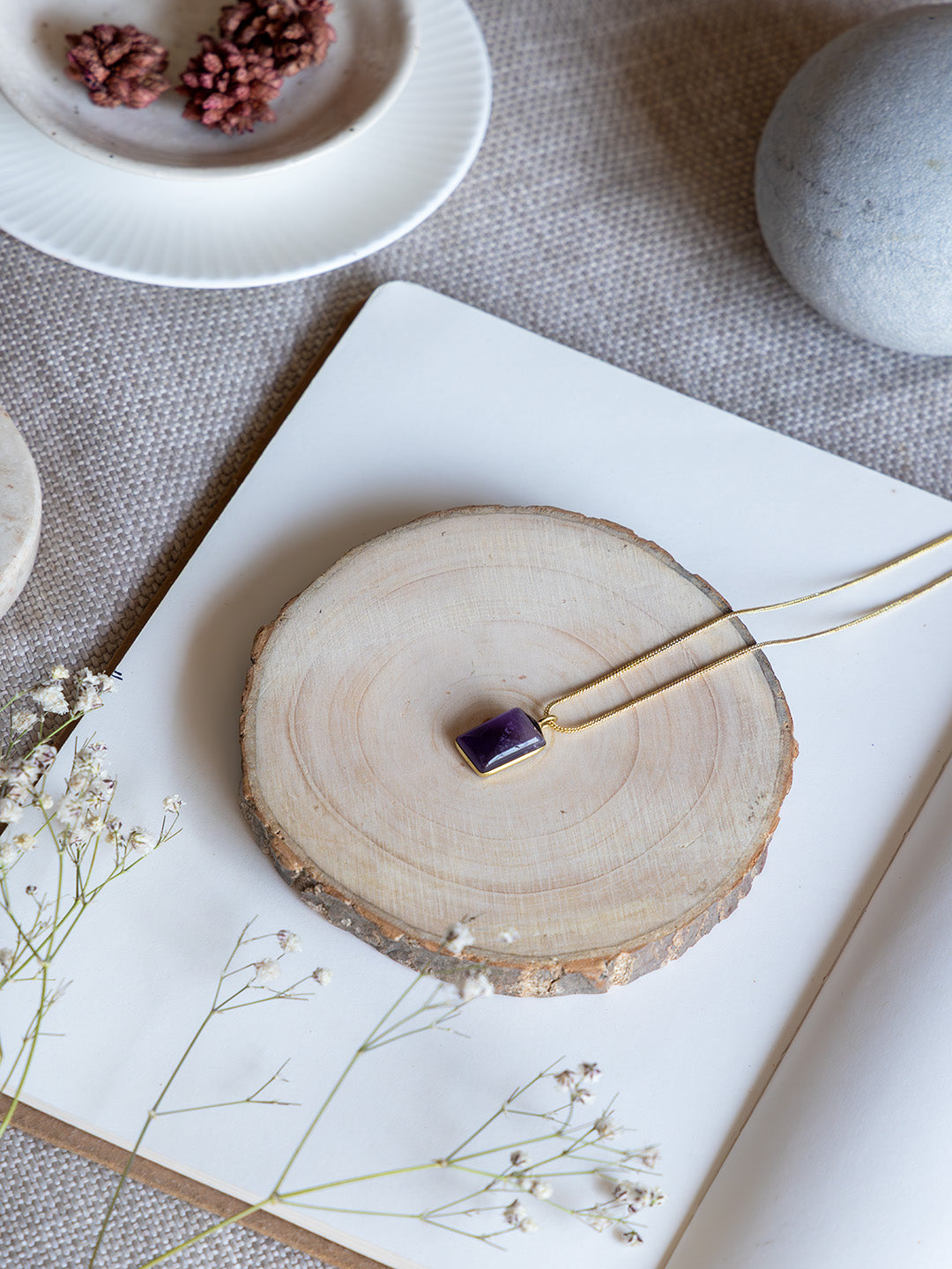 Gold necklace with a purple pendant on a wooden coaster with flowers and stones in the background