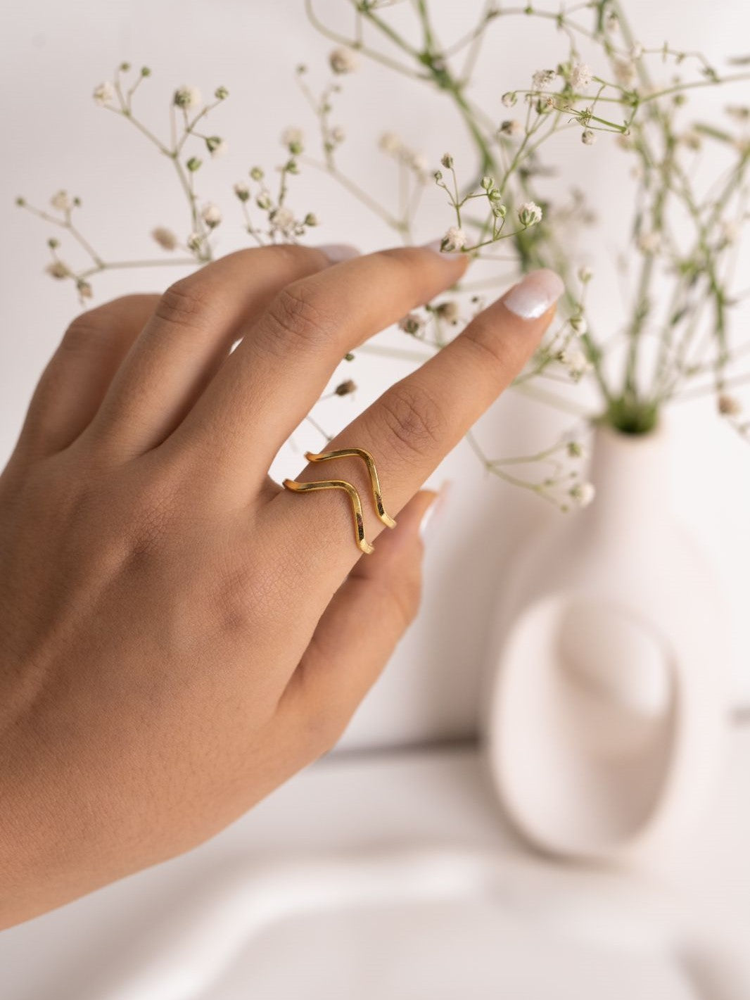 A person's hand wearing a V-shaped brass adjustable ring, with a blurred background featuring a white vase and small flowers.