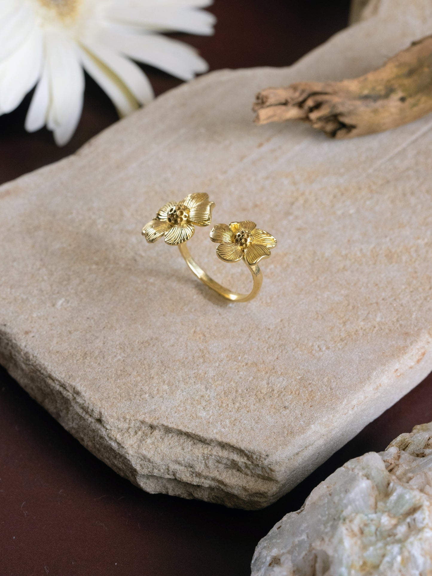 Gold ring with floral design on a stone surface with white flowers and driftwood.