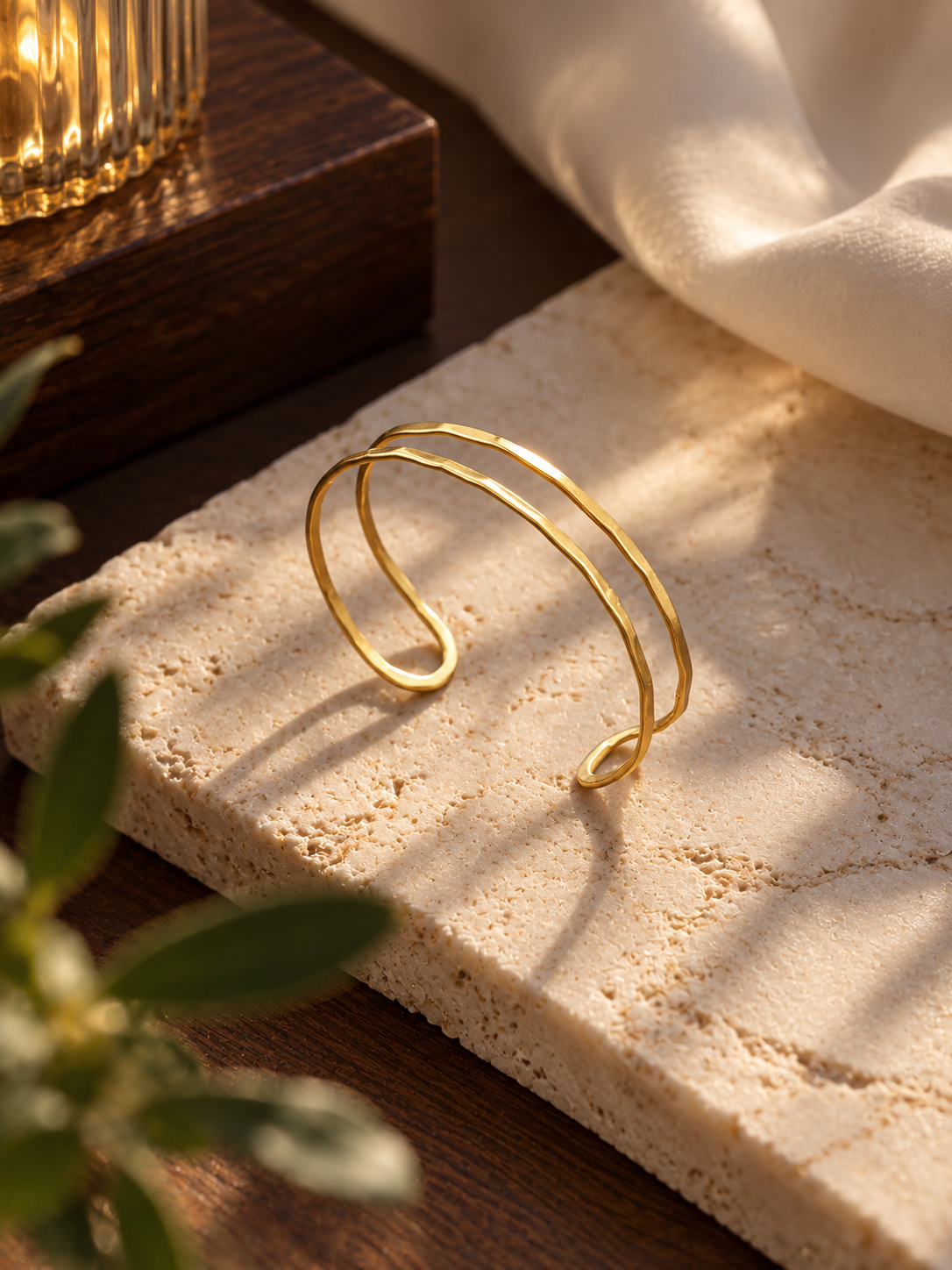 A delicate gold-toned double-band cuff bracelet resting on a textured beige stone slab, with warm sunlight casting shadows, a dark wooden surface, draped cream fabric, and blurred green leaves in the foreground.