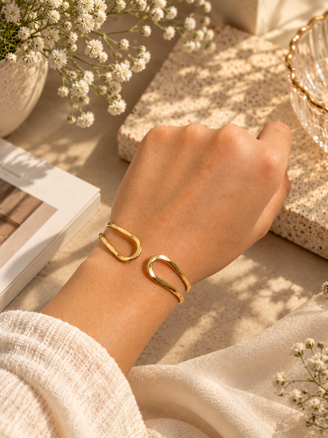 A person's wrist wearing a pair of open cuff bracelets in polished yellow gold, styled with neutral-toned books, dried flowers, and soft fabric in warm sunlight.