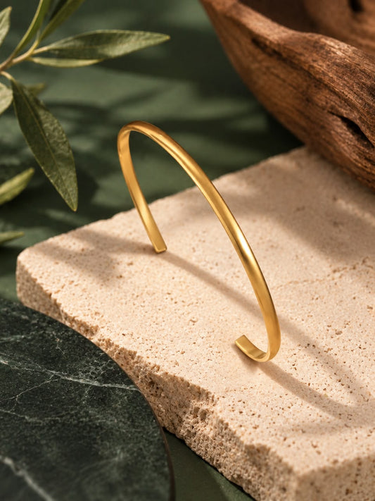 A polished gold open cuff bracelet displayed on a textured beige stone slab with green leaves and a dark wood bowl in the background.