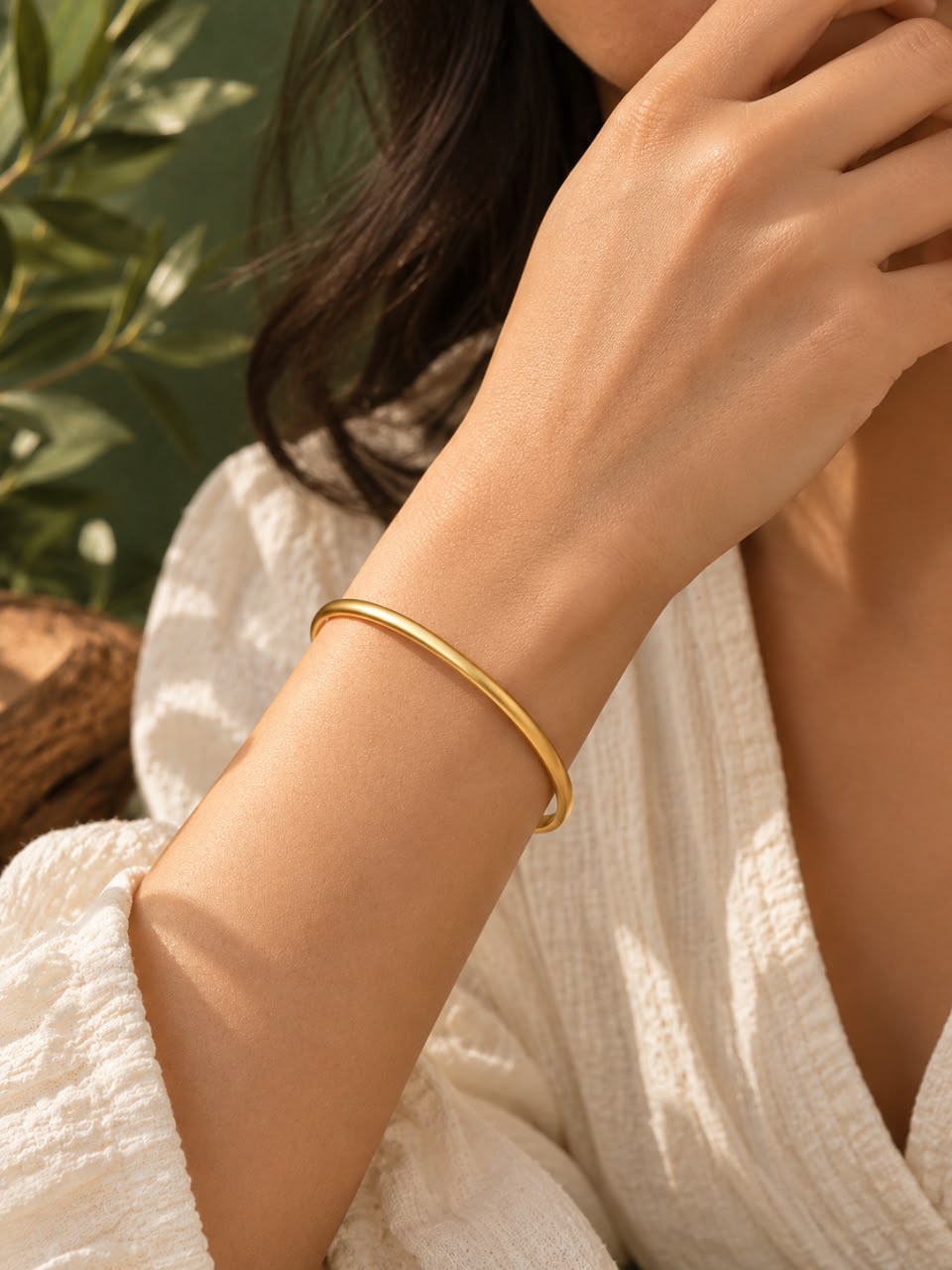 A close-up of a woman's wrist wearing a thin, polished gold bangle bracelet, styled with a textured white top and soft-focus green foliage in the background.