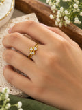 A close-up of a hand wearing a delicate gold ring with a small three-petal flower design set with clear gemstones, styled with baby's breath flowers on a textured stone surface.