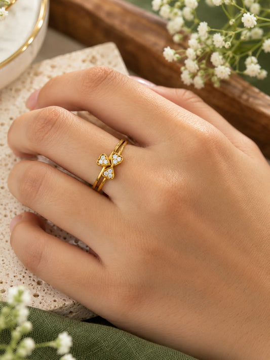 A close-up of a hand wearing a delicate gold ring with a small three-petal flower design set with clear gemstones, styled with baby's breath flowers on a textured stone surface.