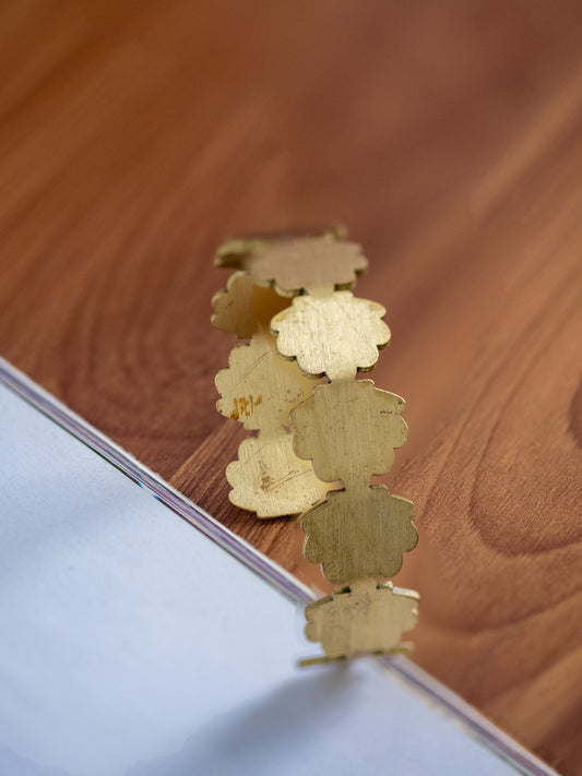 A gold-toned metal cuff bracelet with scalloped leaf-like cutouts, resting on a wooden tabletop next to a white surface.