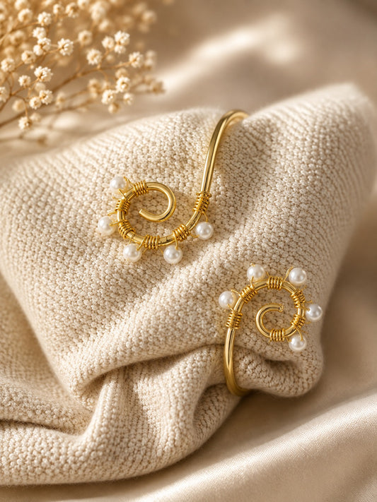 Two gold spiral cuff bracelets with small white pearl accents, displayed on a textured cream knit fabric with dried floral decor in the corner.